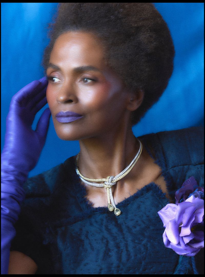 Glamorous studio portrait of a Black woman with purple lipstick and gloves, wearing a jeweled knot necklace against a blue backdrop.