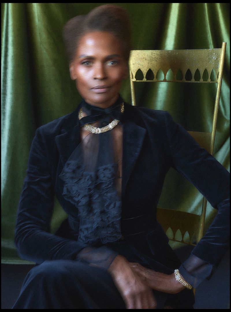 Elegant African-American woman in black velvet blazer and gold jewelry sits in golden chair against an olive green fabric backdrop.