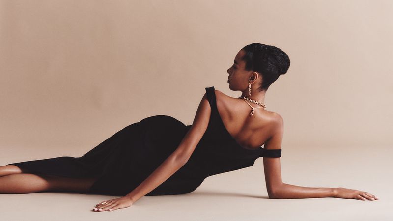 Model Kate Xavier reclining in an off-the-shoulder black dress and gold chain jewelry against a warm beige seamless studio backdrop.