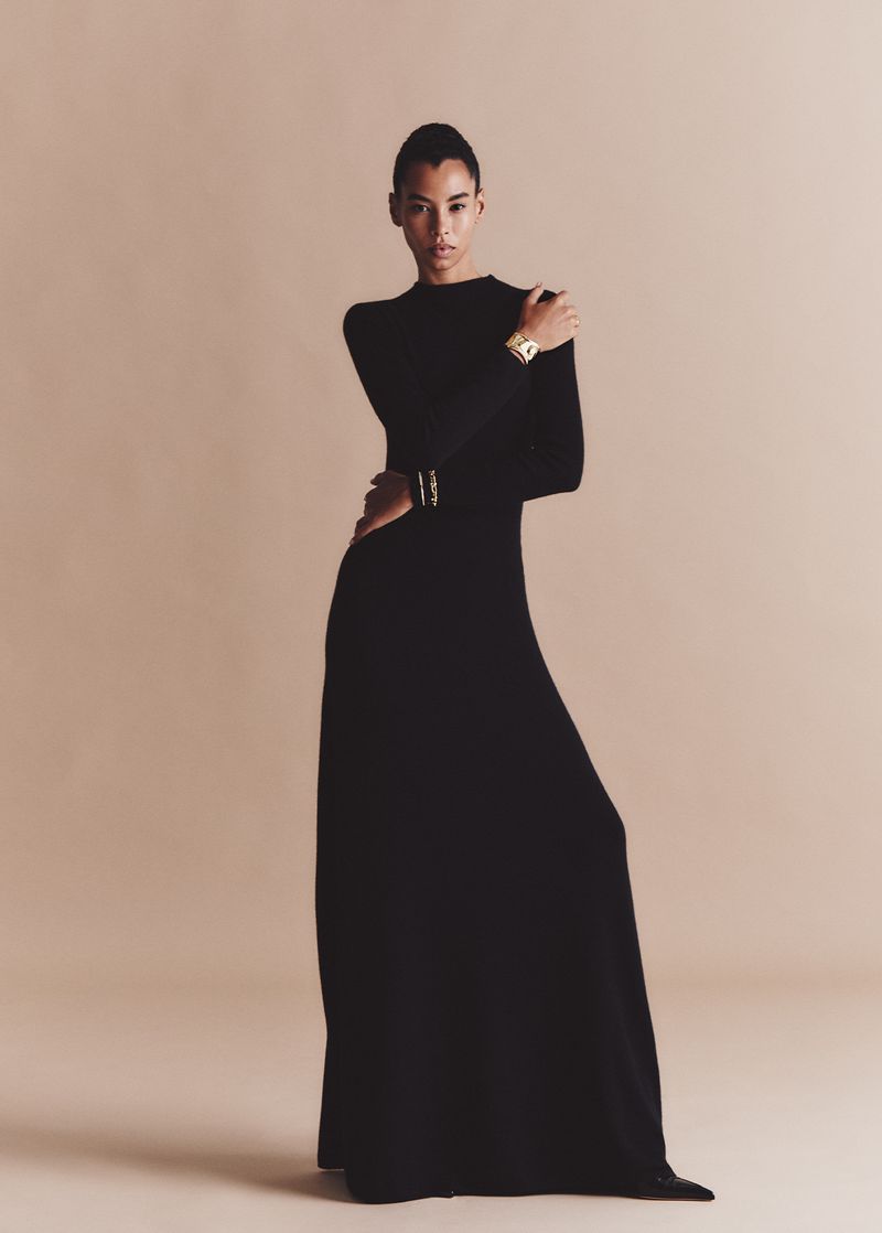 Elegant woman in a long-sleeved black gown, standing in a full shot against a neutral background, with gold bracelets accenting her wrists.