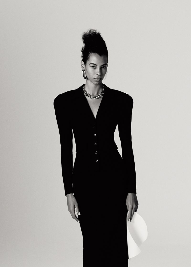 Monochrome fashion portrait of a woman in a black buttoned dress with strong shoulder pads, statement jewelry, and holding a white hat.