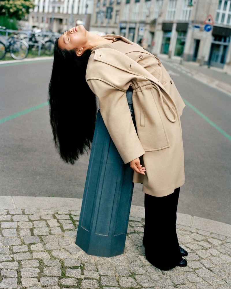 A woman with long dark hair in a beige jacket leans against a blue bollard on a cobblestone street with buildings in the background.