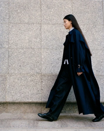 Akari Higashi walks in profile wearing a long oversized navy trench coat and dark trousers against a speckled stone wall.