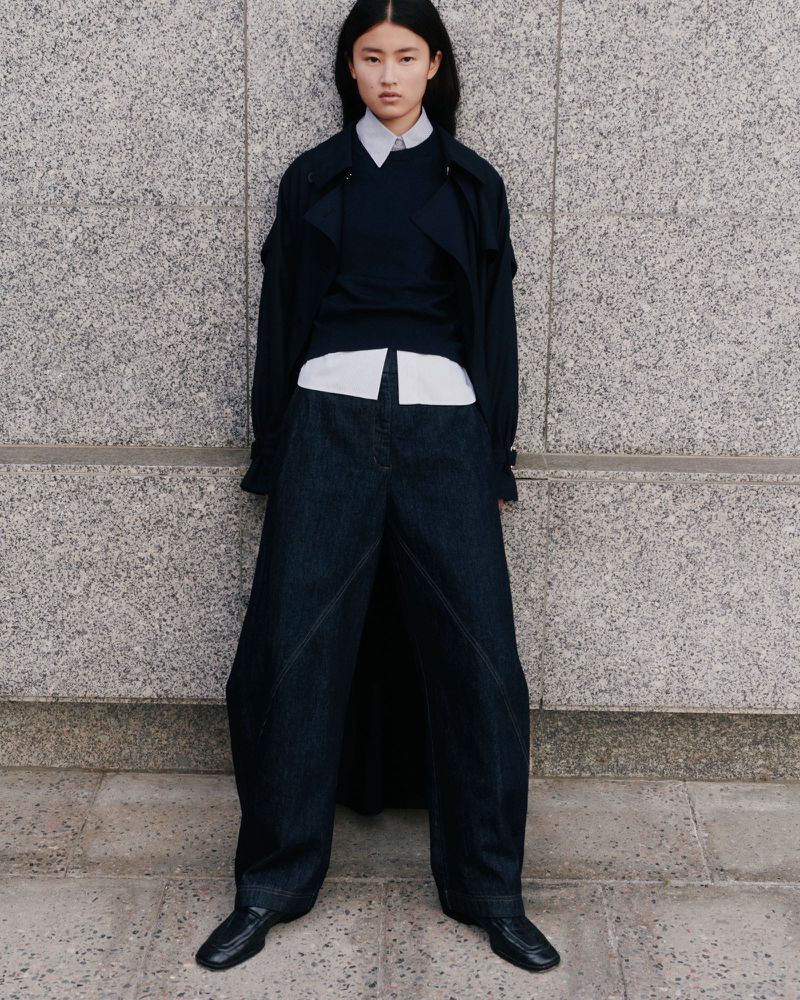An Asian woman stands in a stone urban setting wearing an oversized blue denim pantsuit with a matching sweater and black leather shoes.