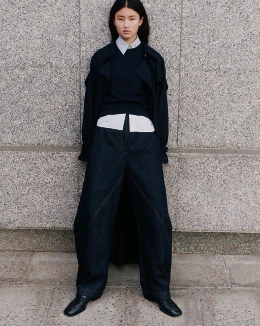 An Asian woman stands in a stone urban setting wearing an oversized blue denim pantsuit with a matching sweater and black leather shoes.