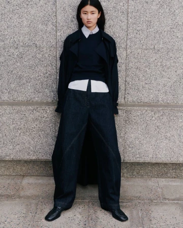 An Asian woman stands in a stone urban setting wearing an oversized blue denim pantsuit with a matching sweater and black leather shoes.
