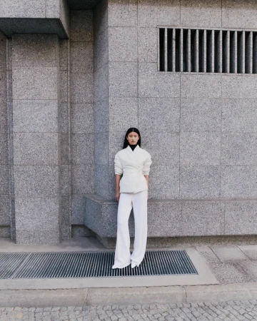 A young Asian woman stands centered against a grey stone wall, wearing a white jacket and flared white pants, posing in an urban setting.