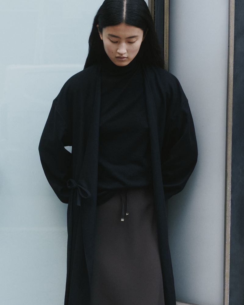 An Asian woman in a black turtleneck and dark skirt stands indoors with natural lighting, modelling a simple, elegant, minimal fashion look.