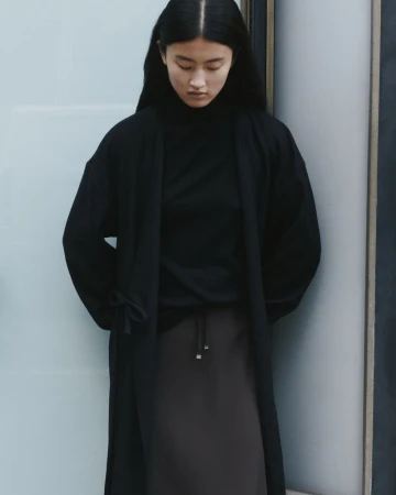 An Asian woman in a black turtleneck and dark skirt stands indoors with natural lighting, modelling a simple, elegant, minimal fashion look.
