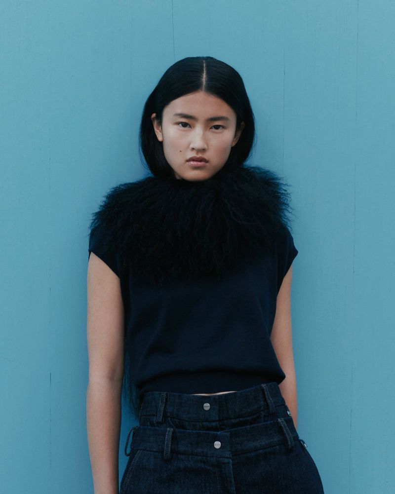 Portrait of a woman with fair skin and dark hair, wearing a black top with feather trim, standing in front of a blue wall.