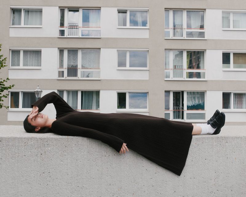 Woman in a brown dress lying on a concrete wall with apartment building in the background, shielding her eyes from the sunlight.