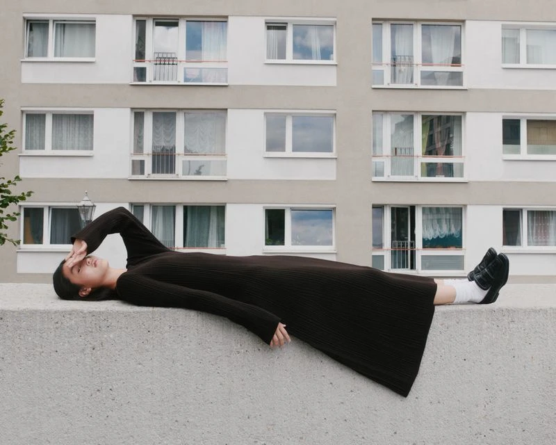 Woman in a brown dress lying on a concrete wall with apartment building in the background, shielding her eyes from the sunlight.