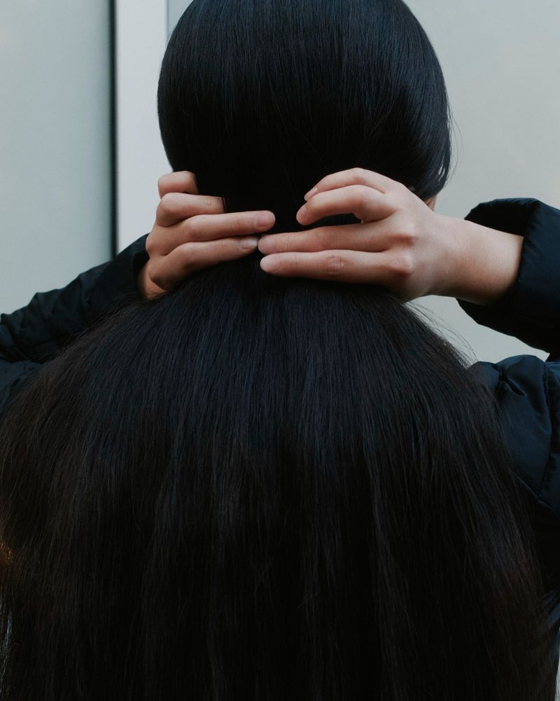 Back view of an adult with long black hair, with hands touching the hair against a neutral background in a close-up shot.