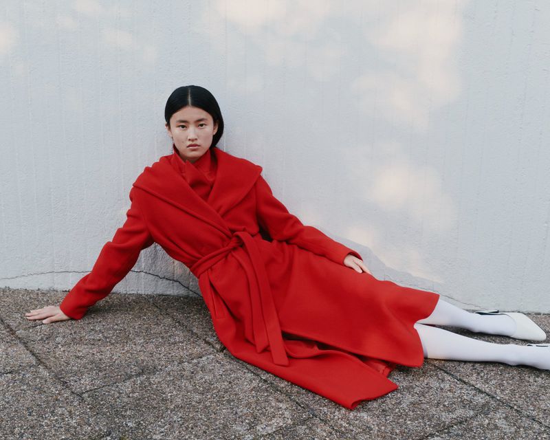 An Asian woman poses in a red coat with a belt, lying on a stone surface next to a white wall with shadow patterns in an outdoor setting.