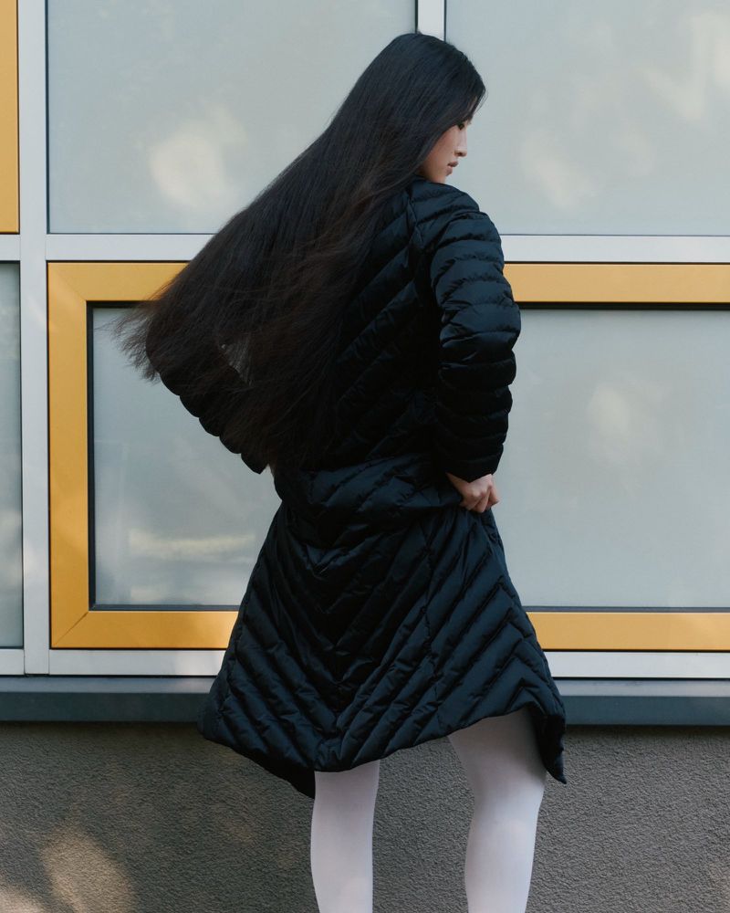 Back view of a woman with long dark hair wearing a black puffer coat and white tights, standing outdoors near a window.