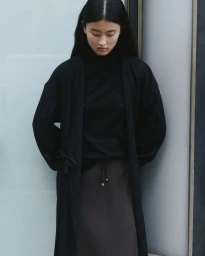 Model Akari Higashi wearing an oversized black coat and high neck top against a frosted glass architectural backdrop.