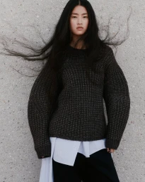 Akari Higashi wearing an oversized dark chunky knit sweater layered over a white shirt with windblown hair against a concrete wall.