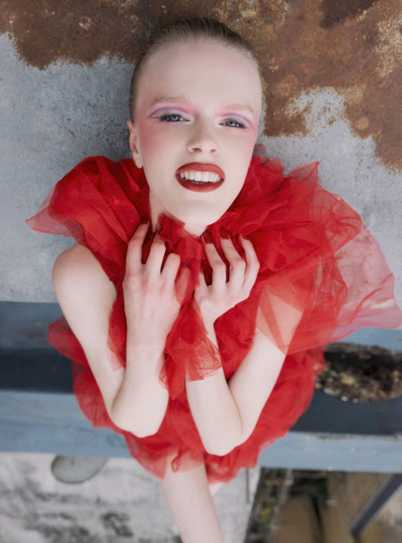 Dramatic fashion portrait: Young woman with red tulle collar, pale skin, pink makeup, and red lipstick poses against concrete backdrop.