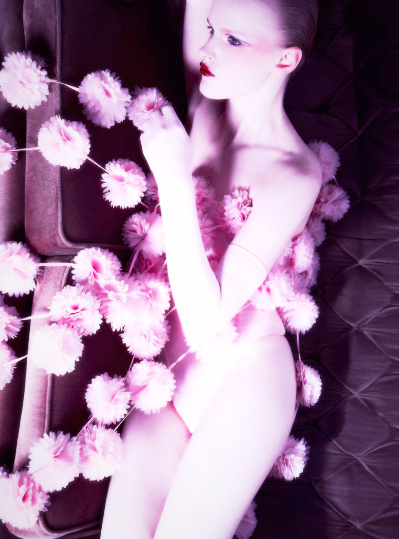 Pale female model with makeup and pink pom poms on a brown quilted surface, laying in a light-filled artistic pose.