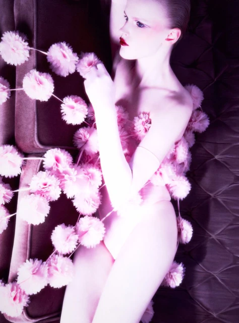 Pale female model with makeup and pink pom poms on a brown quilted surface, laying in a light-filled artistic pose.