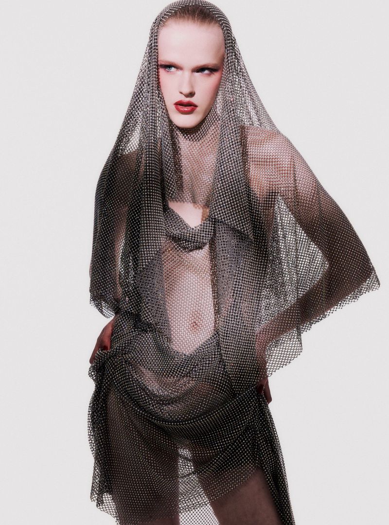 Adult male fashion model poses in a draped silver mesh dress and head covering in a studio with soft, neutral lighting.