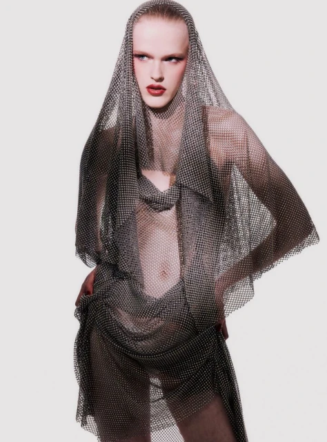 Adult male fashion model poses in a draped silver mesh dress and head covering in a studio with soft, neutral lighting.