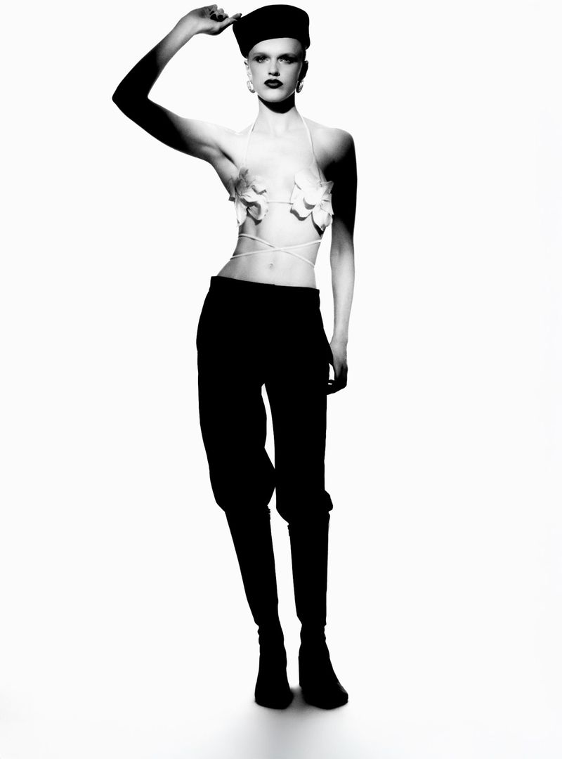 A woman in a fashion forward pose stands in a photo studio wearing a black cap, black pants, and a floral top, all shot in black and white.
