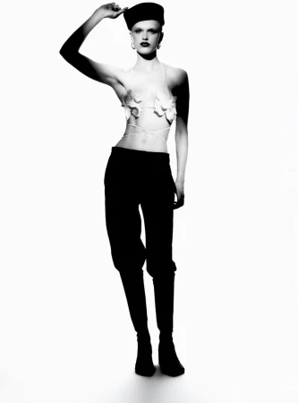 A woman in a fashion forward pose stands in a photo studio wearing a black cap, black pants, and a floral top, all shot in black and white.