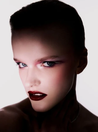 Close-up portrait of a fair-skinned woman with dark hair, striking pink eye makeup, and dark red lipstick against a bright background.