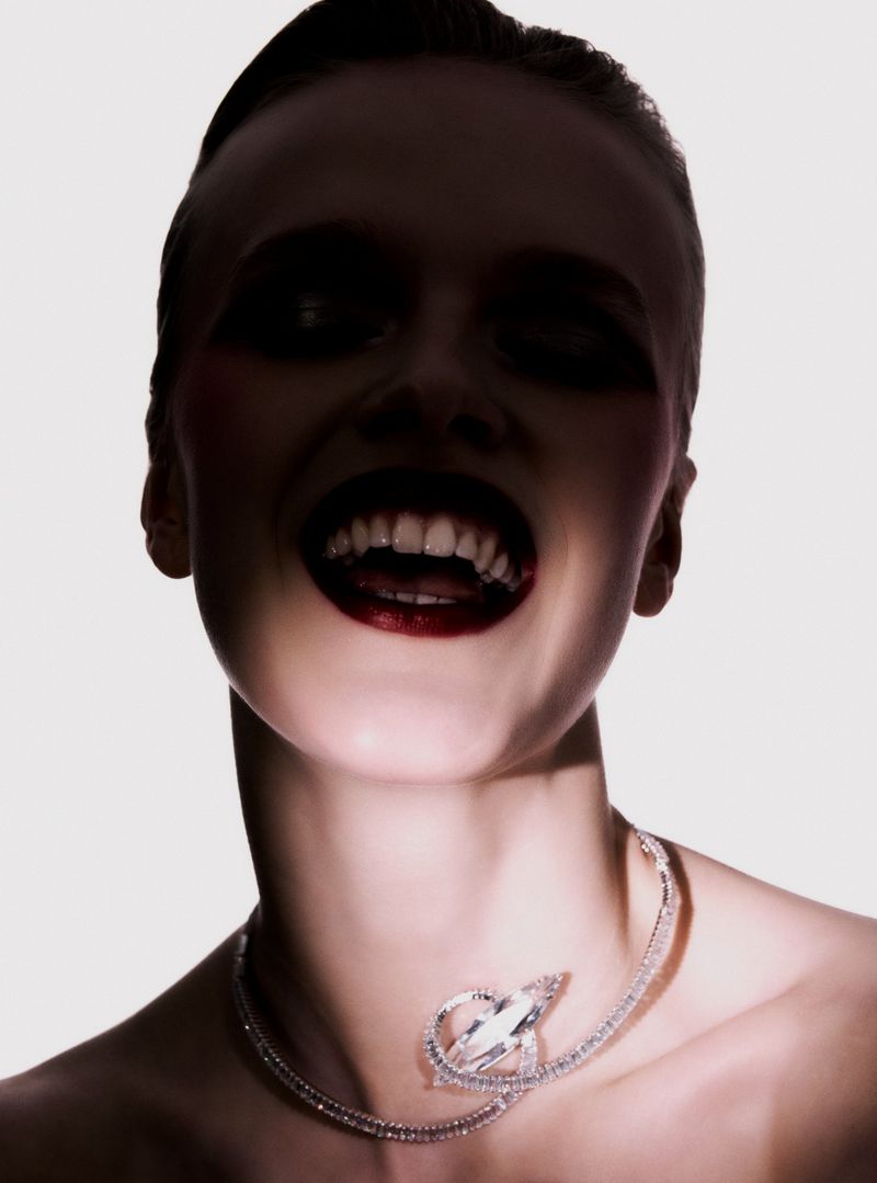 Close-up portrait of a fair-skinned adult woman with dark makeup and a jeweled necklace against a bright white background.