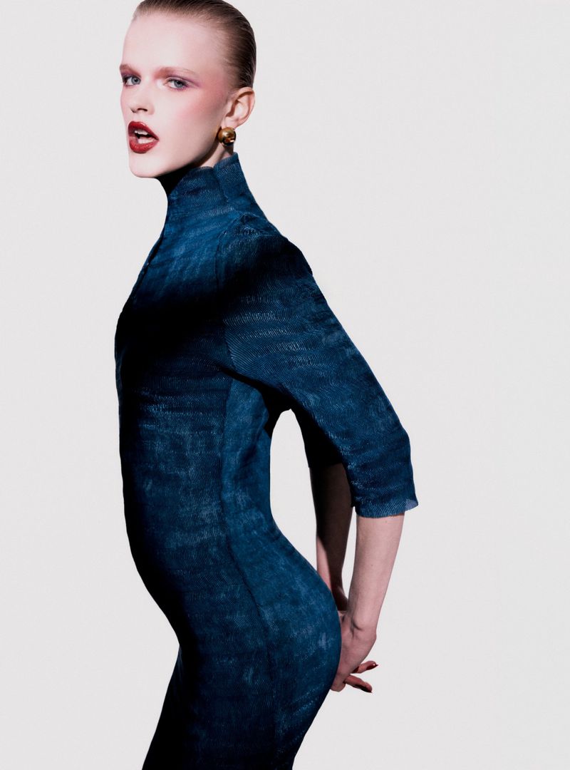 Fashion shot of a fair-skinned adult female model in a form-fitting denim dress, red lipstick and gold earrings against a white background.
