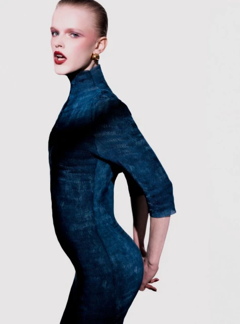 Fashion shot of a fair-skinned adult female model in a form-fitting denim dress, red lipstick and gold earrings against a white background.