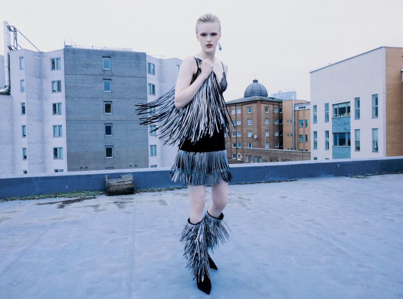 A fair-skinned woman poses on a rooftop in a black dress with silver fringe and matching boots, showcasing a fashion editorial look.