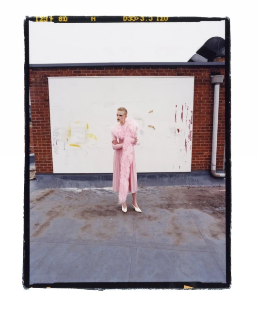 Fashion shot of a blonde adult woman wearing a pink dress and feather boa standing on a rooftop with a brick wall background.