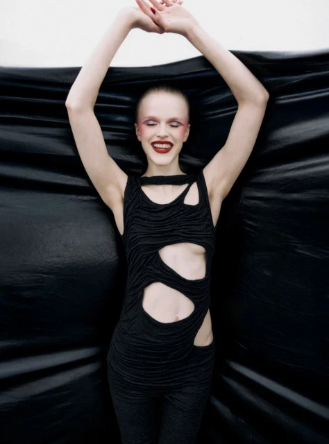 Adult female fashion model poses in a studio wearing a black cut-out top and pants against a black draped background with arms raised.