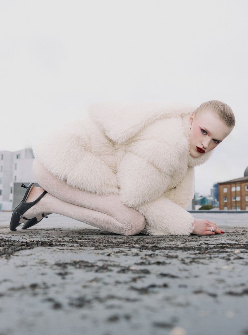 Model Hannah Motler crouching on an urban rooftop wearing a plush cream faux fur jacket, matching tights, and black slingback heels.