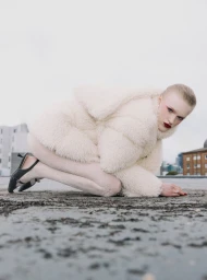 Model Hannah Motler crouching on an urban rooftop wearing a plush cream faux fur jacket, matching tights, and black slingback heels.