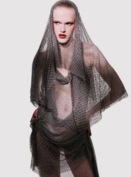 Model Hannah Motler wearing a draped chainmail mesh hooded garment against a light studio backdrop.