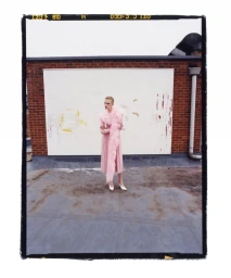 Model Hannah Motler posing in a pale pink feathered dress and white heels on a rooftop against a brick wall and white billboard.