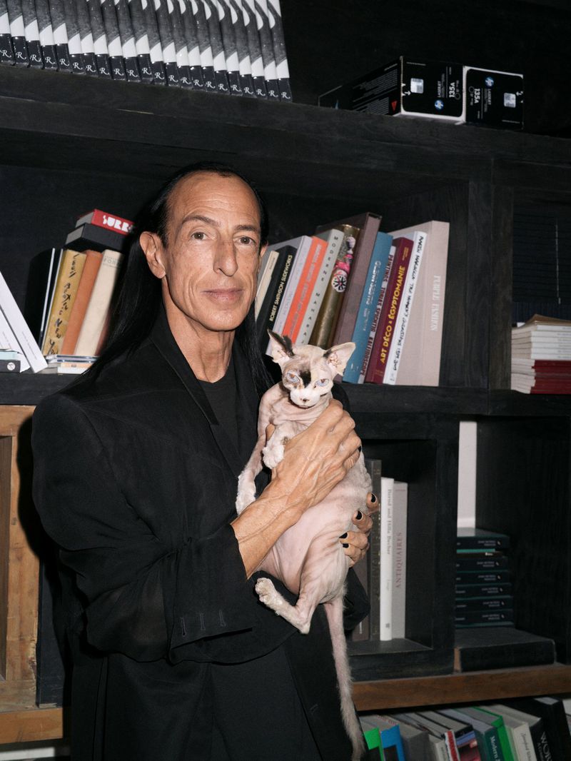 Man poses with a hairless cat indoors. The man wears a black jacket, has long dark hair, and stands near a bookshelf filled with books.