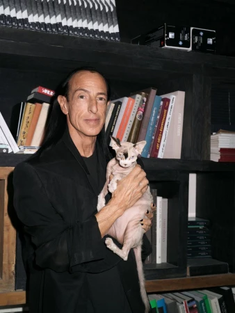 Man poses with a hairless cat indoors. The man wears a black jacket, has long dark hair, and stands near a bookshelf filled with books.