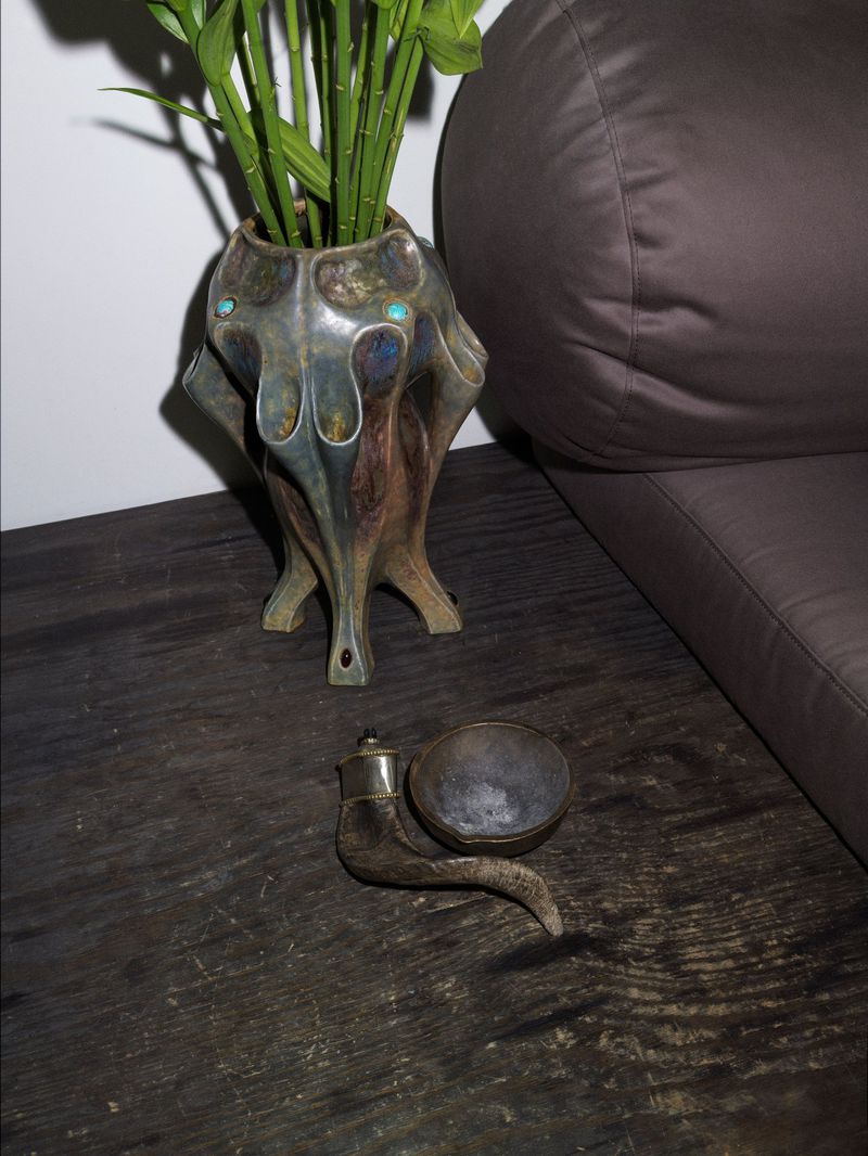 Still life of bamboo shoots in a decorative vase, a smoking pipe, and a dark upholstered couch against a white wall.