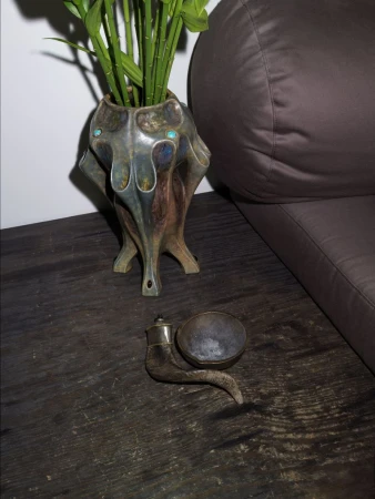 Still life of bamboo shoots in a decorative vase, a smoking pipe, and a dark upholstered couch against a white wall.