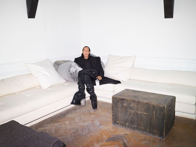 A stylish adult male is sitting on a white couch in a studio with hardwood floors, wearing all black and tall black boots with embellishments.