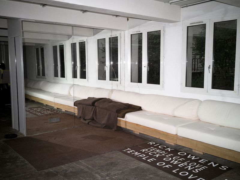 Indoor interior featuring a white modular sofa, windows, a mirror, and a brown rug with the text 'Rick Owens Temple of Love'.