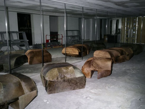 Indoor space featuring multiple upholstered benches with light and dark brown fabric under reflective mirrors, with metal structures in the background.