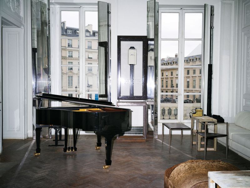 A black grand piano sits in a light-filled room with large windows looking out onto a city street with historic buildings.