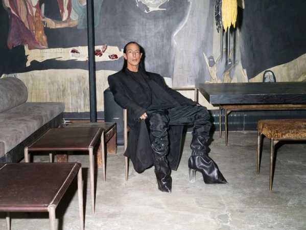 Person wearing a black coat and shiny black boots sitting on a chair indoors with artwork and modern furniture in the background.