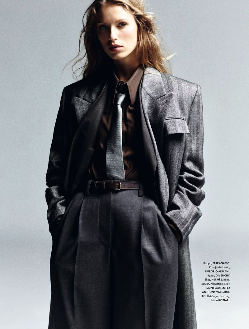 Fashion editorial shot of a woman with long blonde hair wearing a gray suit with a brown shirt and tie in a photo studio.