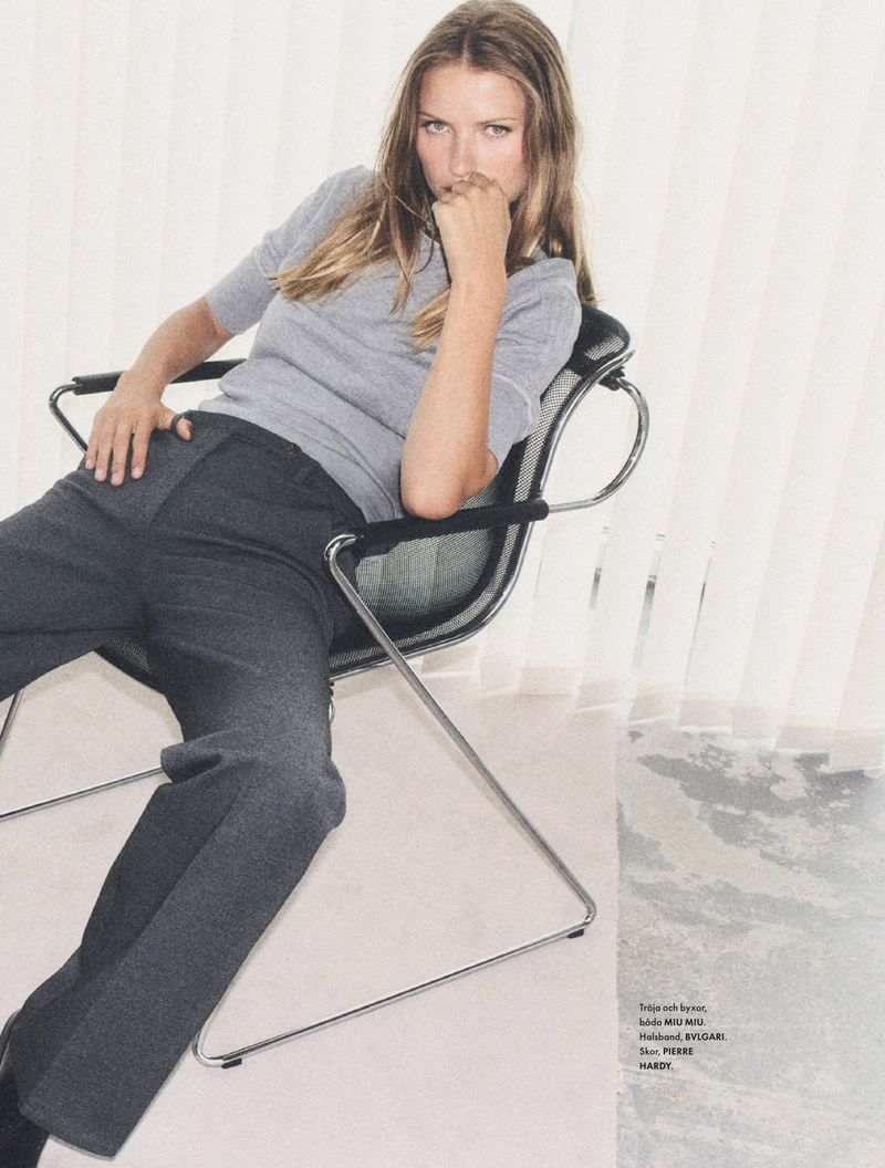A young adult woman is seated on a chair in a studio, wearing a gray sweater and gray slacks, with a window visible in the background.